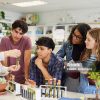 Mature science teacher showing sample in test tube to a group of multiethnic students during classroom activity. High school guys and girls gathered around table to observe biology experiment involving plant specimen. Group of young college classmates working on science project at school.