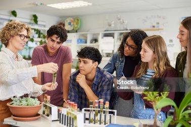Mature science teacher showing sample in test tube to a group of multiethnic students during classroom activity. High school guys and girls gathered around table to observe biology experiment involving plant specimen. Group of young college classmates working on science project at school.
