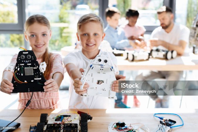 Talented children. Charming joyful brother and sister standing near the working table during their robotics workshop and showing their robot models, having constructed them themselves