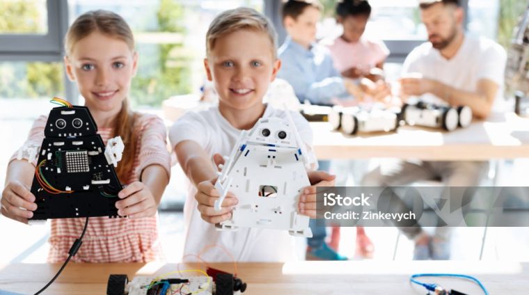 Talented children. Charming joyful brother and sister standing near the working table during their robotics workshop and showing their robot models, having constructed them themselves